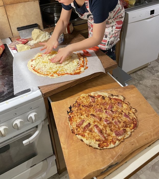 Person förbereder pizza bredvid en bakad pizza på ett köksbord. Matlagning, hemgjord mat, köksmiljö.