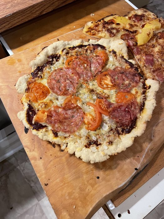 Hembakt pizza på träskärbräda med salami, tomat, ost, ananas och synliga brända delar.