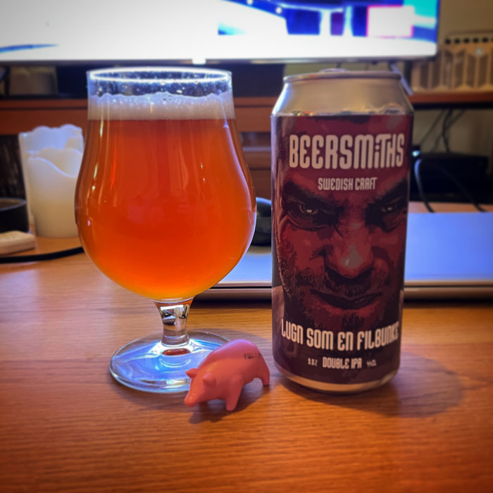 Ölglas fyllt med öl, burk märkt "Beersmiths", liten rosa plastgris, datorskärm i bakgrunden.
