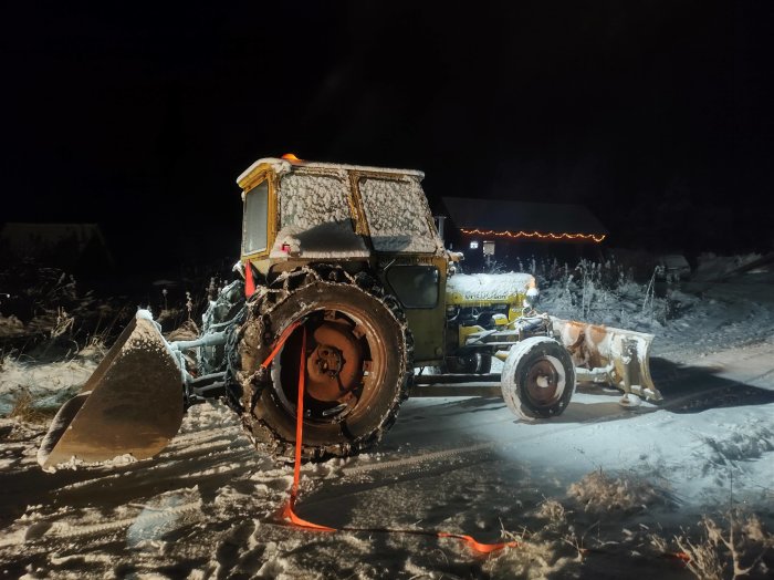 Gammal traktor täckt med snö, kväll, julbelysning i bakgrunden, leriga hjul, snöig miljö.