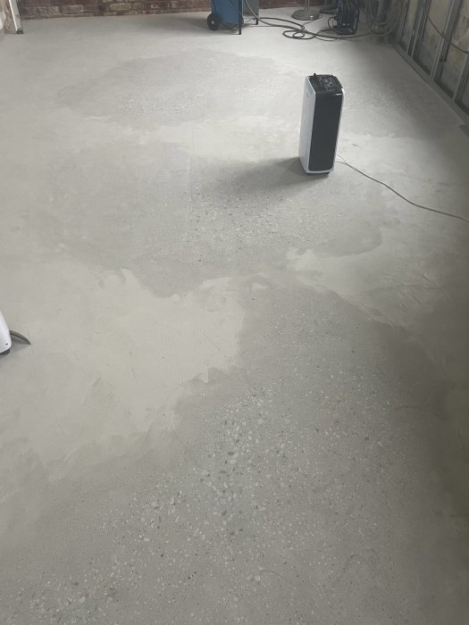 Ett avfuktare i ett outrett rum med slät betonggolv och tegelvägg. Kabel löper nerifrån maskinen.