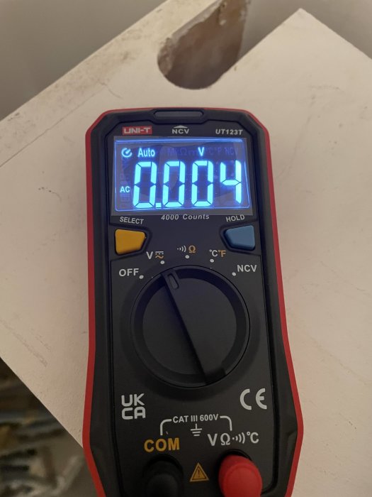 Digital multimeter visar 0.004 i en AC-spänningsmätning, hållen framför oren yta.