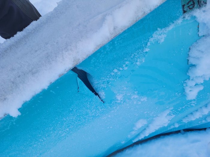 Skadad blå bil med snötäckt vindruta och kaross, troligen efter olycka eller storm.