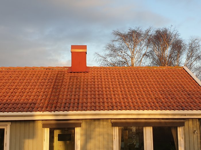 Ett hus med rött tak och skorsten i solnedgångens ljus, träd i bakgrunden.