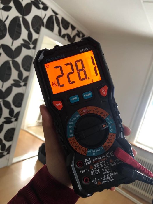 Hand håller digital multimeter visar spänning 228.1 volt, bakgrund med bladmönstrad tapet.