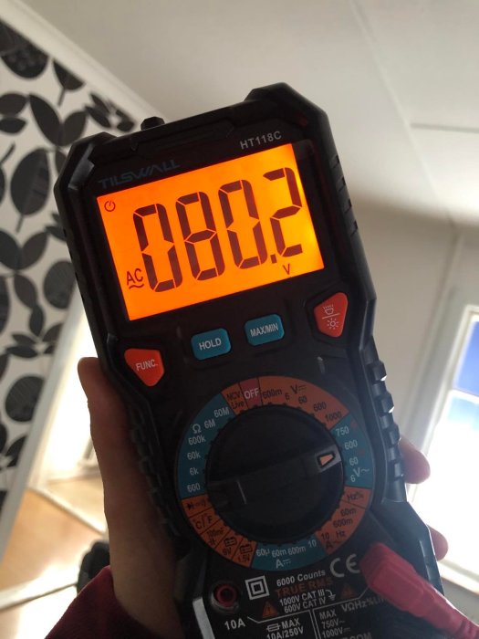 Digital multimeter hålls upp, visar 0.02 volt, i bakgrunden syns en vägg med bladmönster.