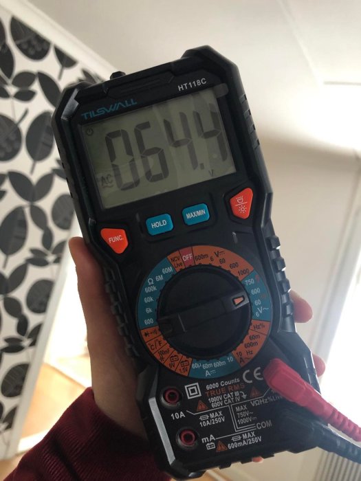 Hand håller en multimeter som visar 06.44 V, mot en bakgrund av en vägg med tapet.