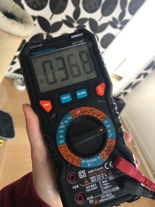 En hand håller en digital multimeter med displayen som visar 036.8 i en inomhusmiljö.