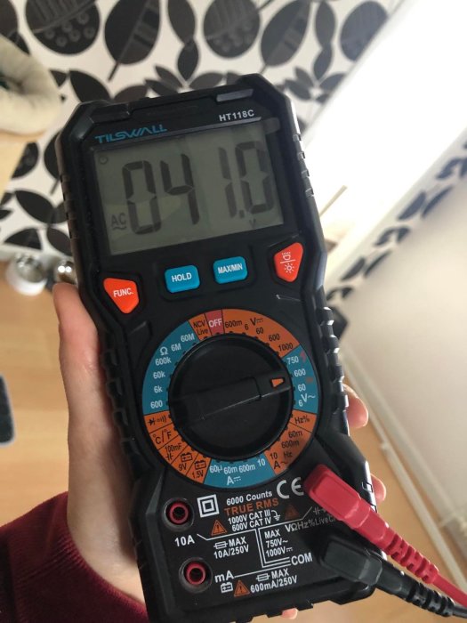 Hand håller en multimeter som visar 0,41 AC volt. Bakgrund med lövmönstrad tapet.