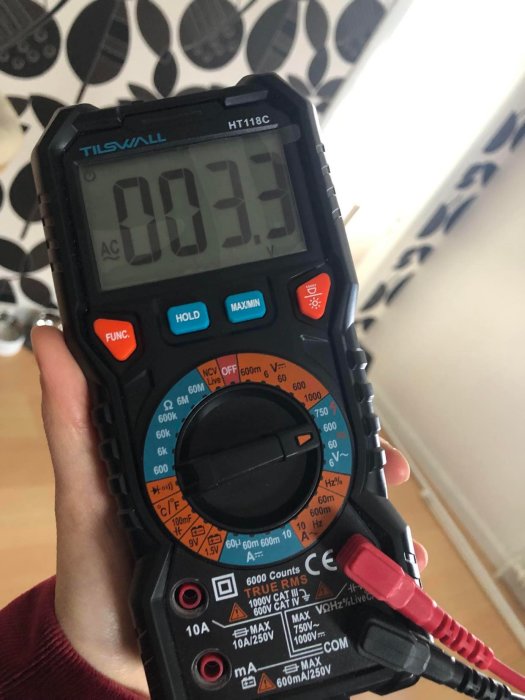 Multimeter visas närbild, mäter spänning, visar 0.33 volt, hålls i hand, bakgrund med mönstrad tapet.