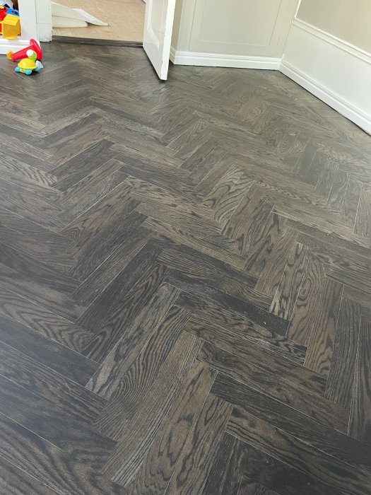 Mörkt klinkergolv med träeffekt, leksak till vänster, vit dörr och vägg, ljusinfiltration.