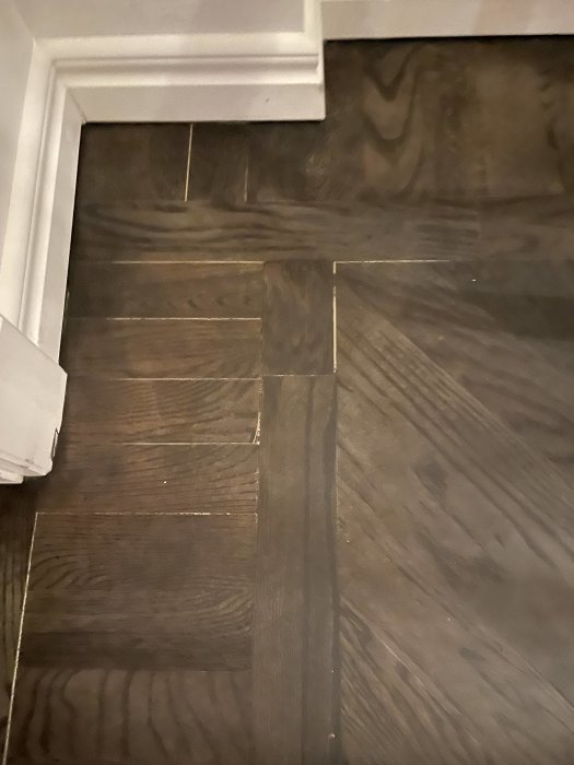 Mörkt trägolv med synliga skarvar, vit dörrkarm nederkant, inomhus, hemmiljö.