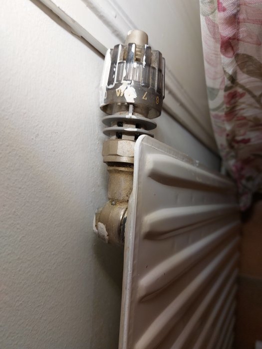 Termostatventil på en radiator, vit vägg, slitna märken, inomhus, rörkoppling, värmeinställning.