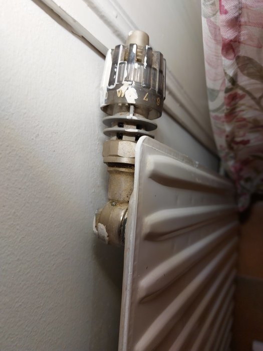 Termostatventil på radiator, sliten med nummer, plast och metall, fastsatt vid vägg, gardinfragment högerkant.