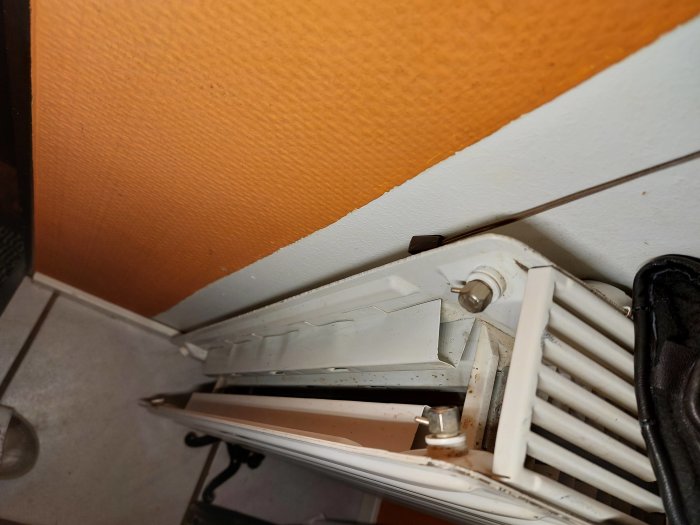 Närbild av vit radiator, orange vägg, vit taklist, golvplattor, oskarp svart föremål, oskarp interiör.