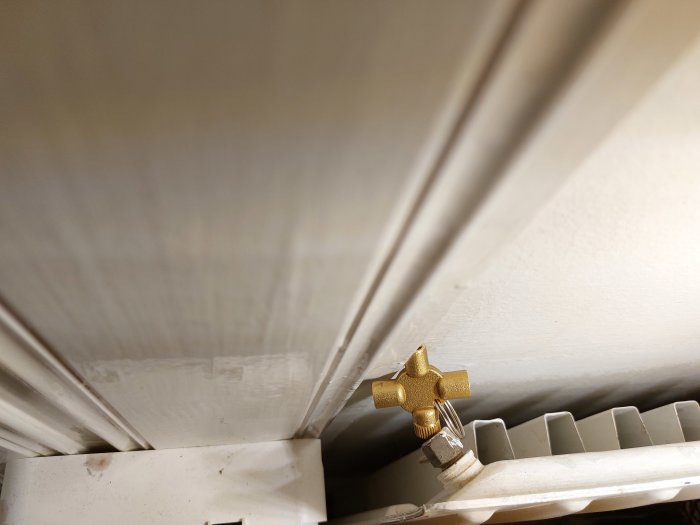 Guldfärgad ventil ovanpå radiator, under ett vitt tak, inomhus, med skuggor och dämpad belysning.