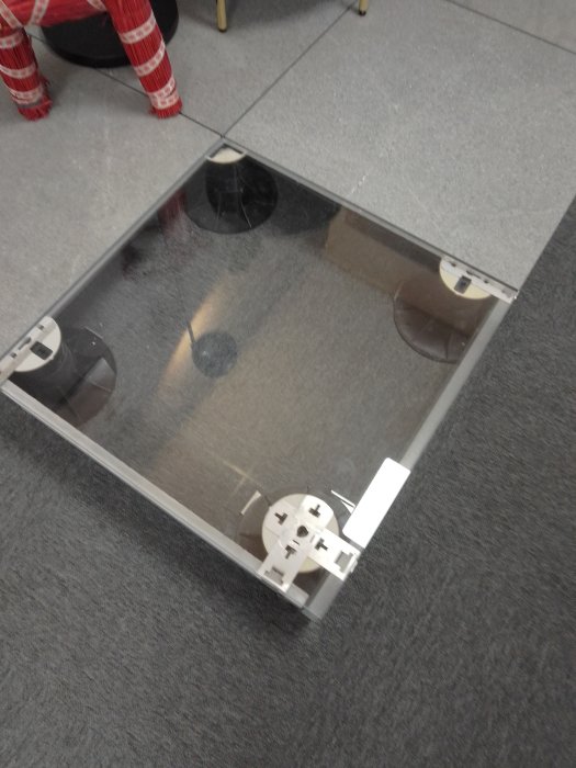 Transparent kvadratiskt bord på grå matta, reflekterande yta, suddiga objekt, designmöbel, samtida kontorsmiljö.