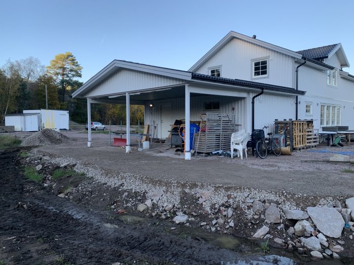 Ett vitt hus med carport under konstruktion, grus, skräp och byggmaterial spridda, klart för landskapsarbete.