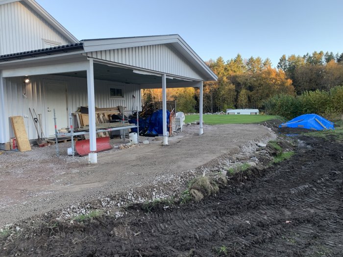 Lantgårdsbyggnad med carport, byggmaterial, grävmaskinsspår, blå presenning, höstträd i bakgrunden, kvällsljus.