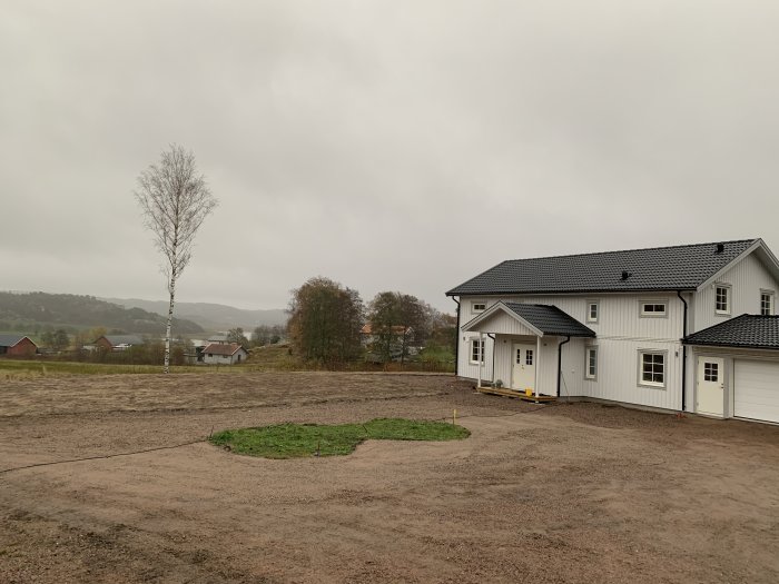 Vitt hus med snedtak, molnig himmel, bar mark, ett ensamt träd, byggnadsarbete pågår, dystrare väder.