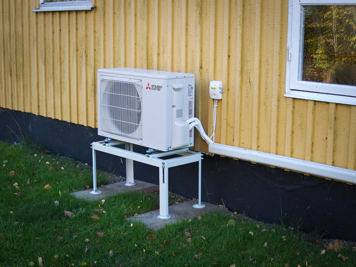 Luftvärmepump installerad utanför gult hus, grön gräsmatta, rör anslutna, fönster, höstlöv.