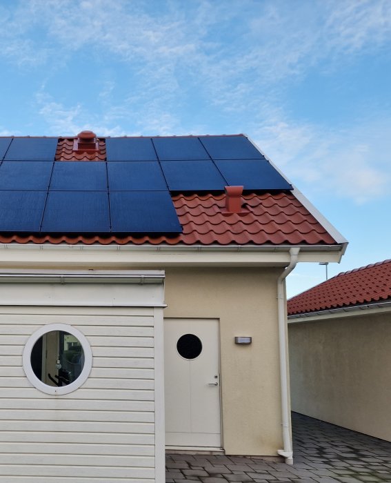 Ett hus med solpaneler på taket, vit fasad, rundat fönster, blå himmel.