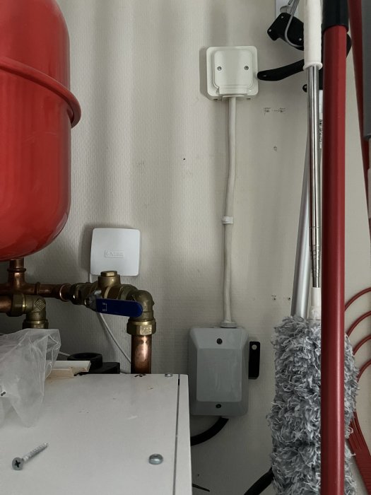 Utrymme med röd expansionstank, kopplingar, elektriska dosor och städmateriel på en randig tapet.