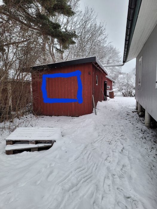 Vinrött skjul, snö, avsmalnande gångväg mellan hus, träbänk, grå vinterdag, blå markering på bilden.