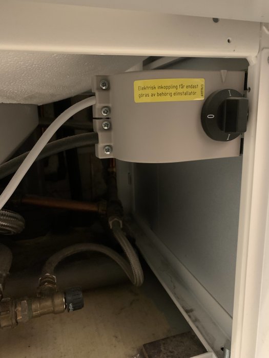 Vattenanslutningar och elektrisk brytare under vit apparat med varningsetikett om behörig installatör.