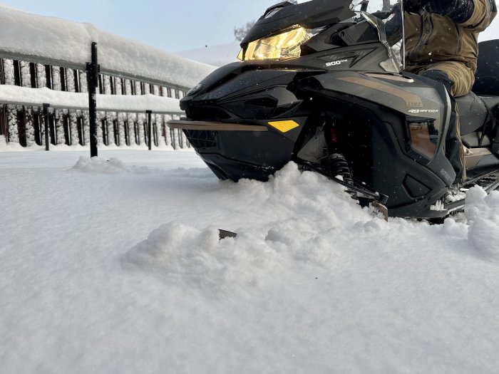 Snöskoter framför snötäckt staket, guldimma framlyse, förare i vinterkläder, fräscht snölandskap.