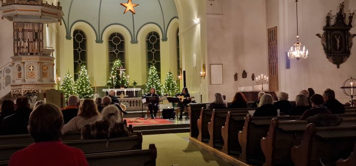 Julkonsert i kyrka med publik och musiker, dekorerade julgranar, stjärna, levande ljus, arkitektoniska detaljer.