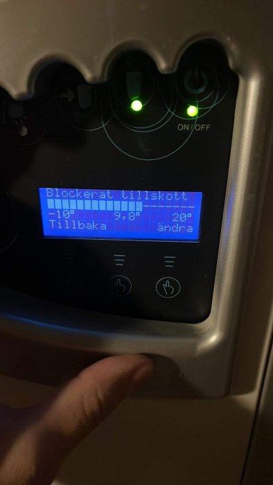 Digital display på vitvaror, fingrar som trycker, text "Blockerat tillskott", indikerar varuinställning eller felmeddelande.