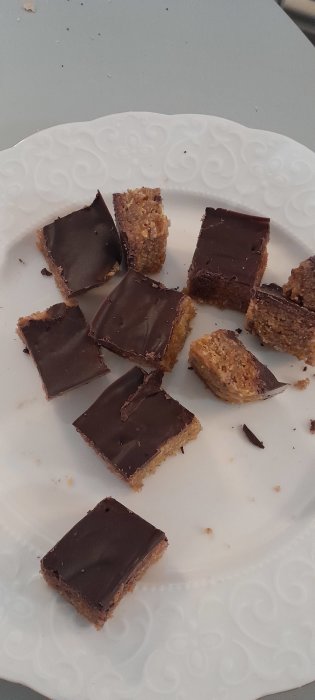 Ett vitt mönstrat fat med små bitar chokladöverdragna havrekakor eller bars. Verkar vara hemmagjorda. Några är halvätna.