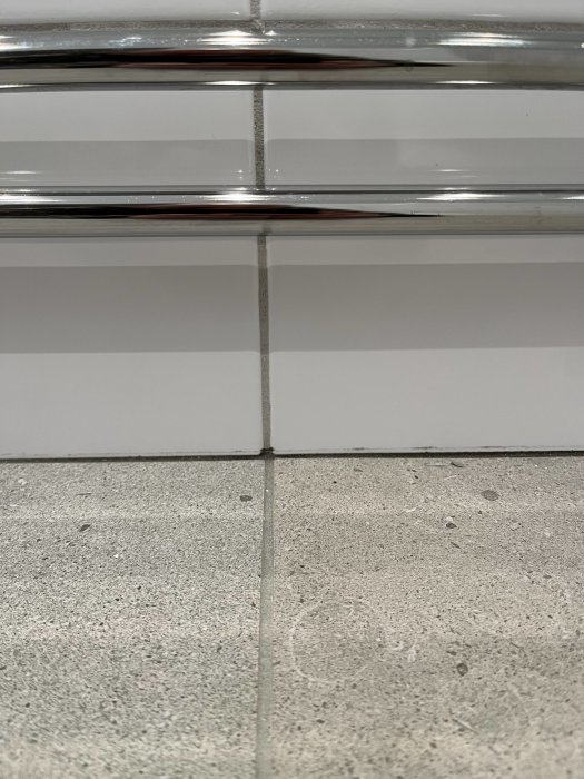 Kromat handdukstork framför vit kakelvägg över grått golv. Vertikal och horisontell linjekonvergens, minimalistiskt, rent.
