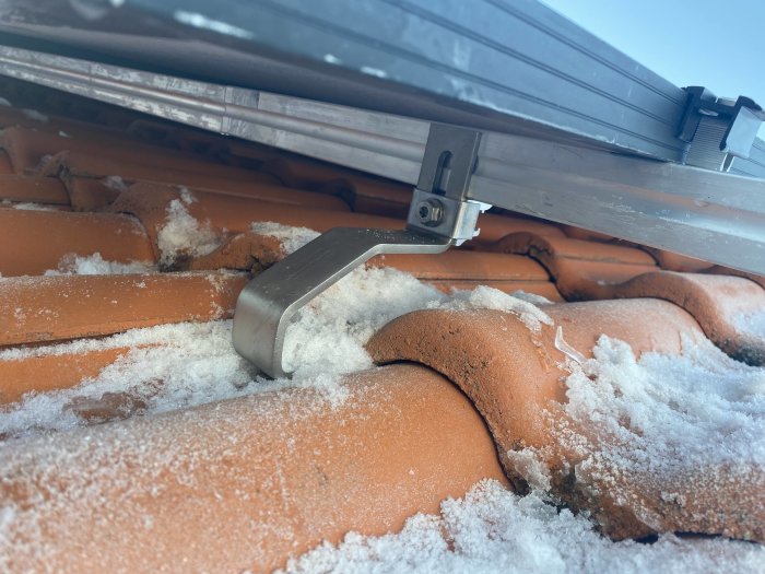 Snö på takpannor med en metallfäste och en del av ett snörasskydd.