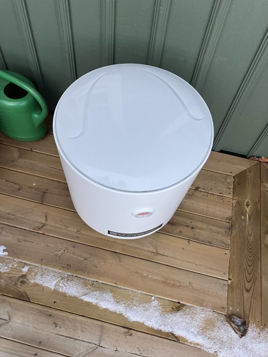 Vit komposthink med lock på träveranda, bredvid en grön vattenkanna, snörest.