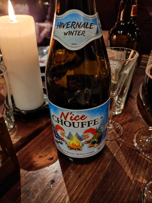 Ölflaska med etikett "N'ice Chouffe", tända ljus, glas, träbord, mysig atmosfär.