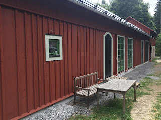 Rött trähus med vita knutar, fönster och dörrar, bänk och bord på grusgång, grönska i bakgrunden.