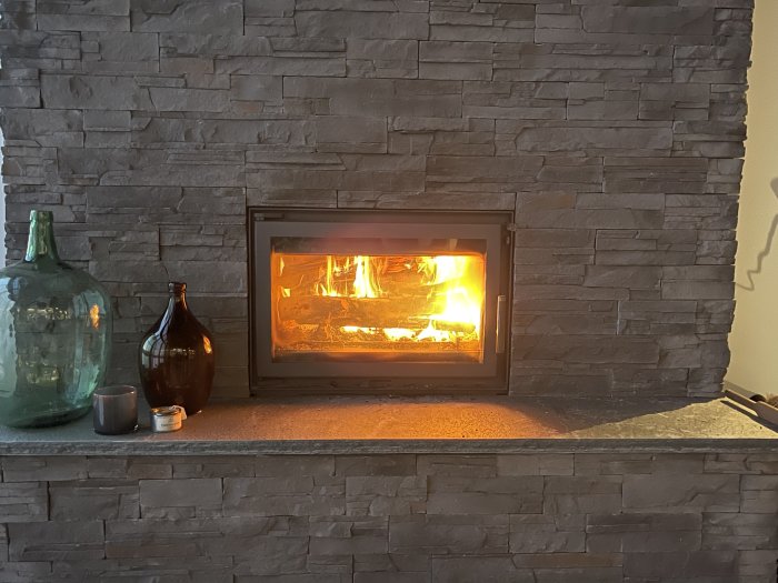 Eld brinner i en modern öppen spis med stengrund, flankerad av dekorativa glasflaskor och ett ljus.