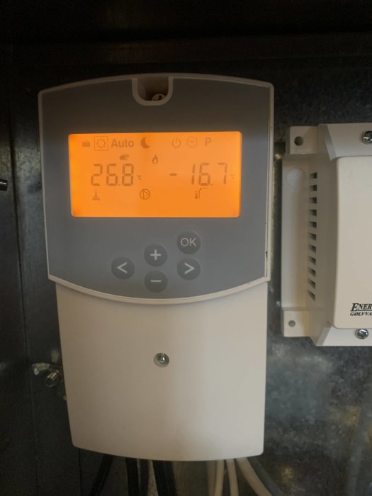 Digital termostat med bakgrundsbelyst display visar inomhus- och utomhustemperatur i Celsius. Monterad på mörk vägg.