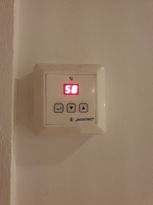 Vit elektronisk enhet på väggen med röda siffror som visar 58, knappar och "acetec" logotyp.