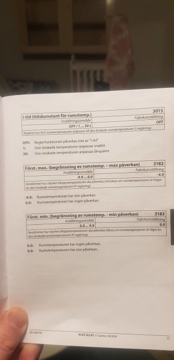 Svensk manual för temperaturreglering, hand håller sidan, inomhusmiljö syns suddigt i bakgrunden.