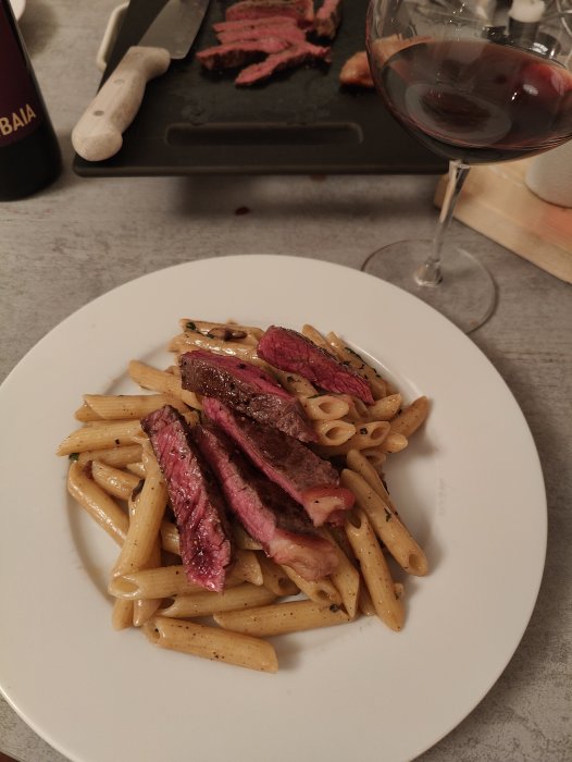 Middagstilläggning med pasta och medium-rare biff, rödvin i bakgrunden, kökskniv och skärbräda.