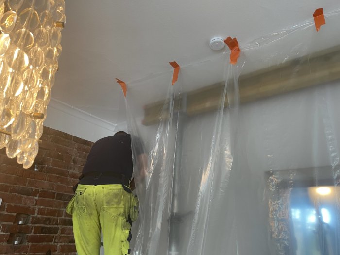 Person i arbetskläder reparerar tak; plastskydd hänger ner; tegelvägg och designlampa synliga.