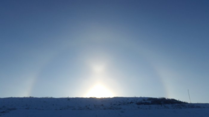 Vinterlandskap, solnedgång, halo-fenomen, solpelare, snöklädd mark, kallt väder, klar himmel, optisk atmosfärisk effekt.