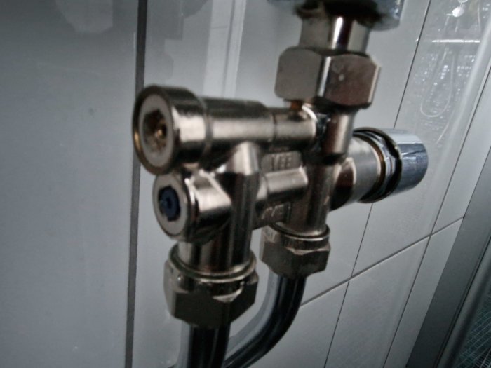 Metalliska vattenanslutningar och ventiler mot kakelvägg, troligen del av dusch eller badrumsinstallation.