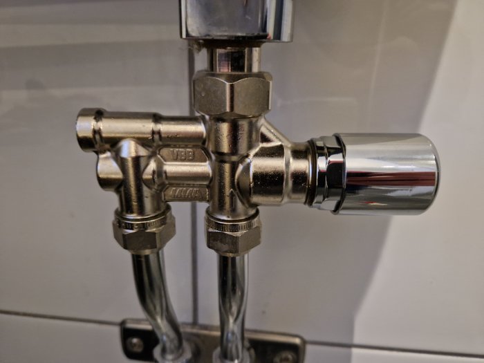 Metallisk toalettspolningsventil med kromfinish och VVS-kopplingar monterad på kakelvägg.