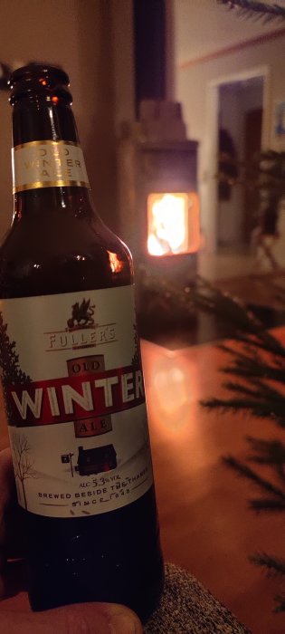 Ölflaska nära kamin, Fullers Winter Ale, mysig inomhusmiljö, lugn atmosfär, mörkt rum, julgran i bakgrunden.