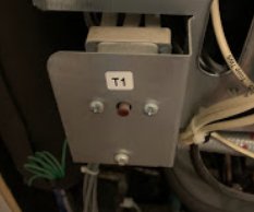 En del av en apparat med etiketten "T1", kablar, en röd lysdiod, och metallkomponenter inneslutna i en mörk miljö.