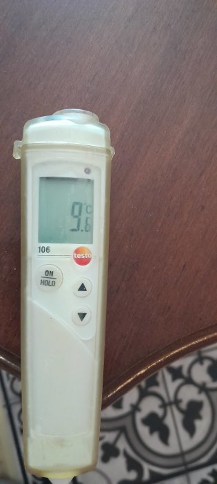 Digital termometer visar 9,6 °C, på tråkig träbakgrund och mönstrat underlag. Verktyget är smutsigt och använd.